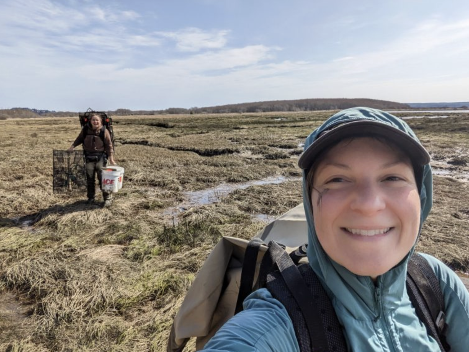 2023GreenCrabStatus_AlexStoteAlexaBrown A selfie of Alex in the field on a coastal marsh with Alexa in the background carrying a bucket and shrimp traps