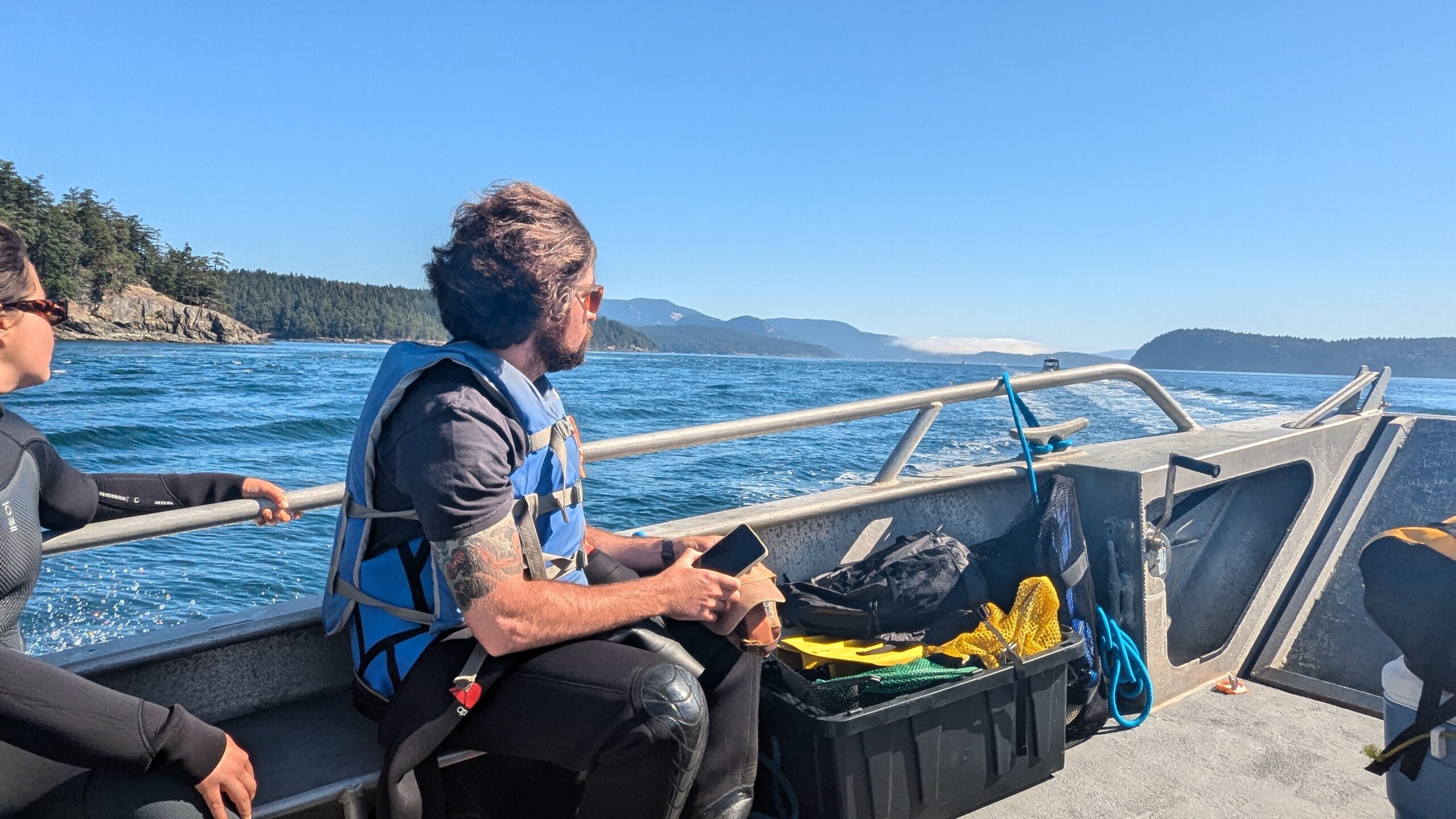 Hobson Boat Walker sits on a seat of a small metal boat. In the background the San Juan Islands can be seen.