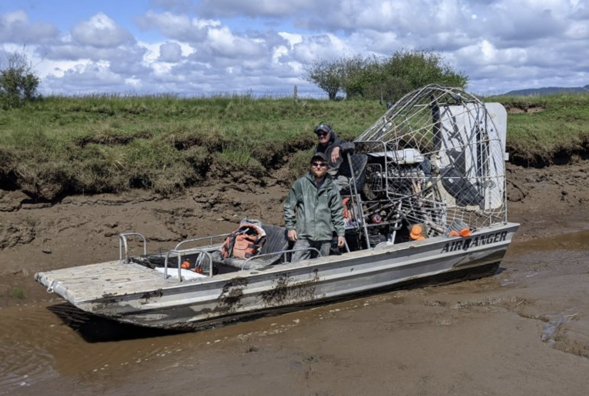 An airboat with two men on it sits in shallow water in a muddy salt marsh channel.<br />
