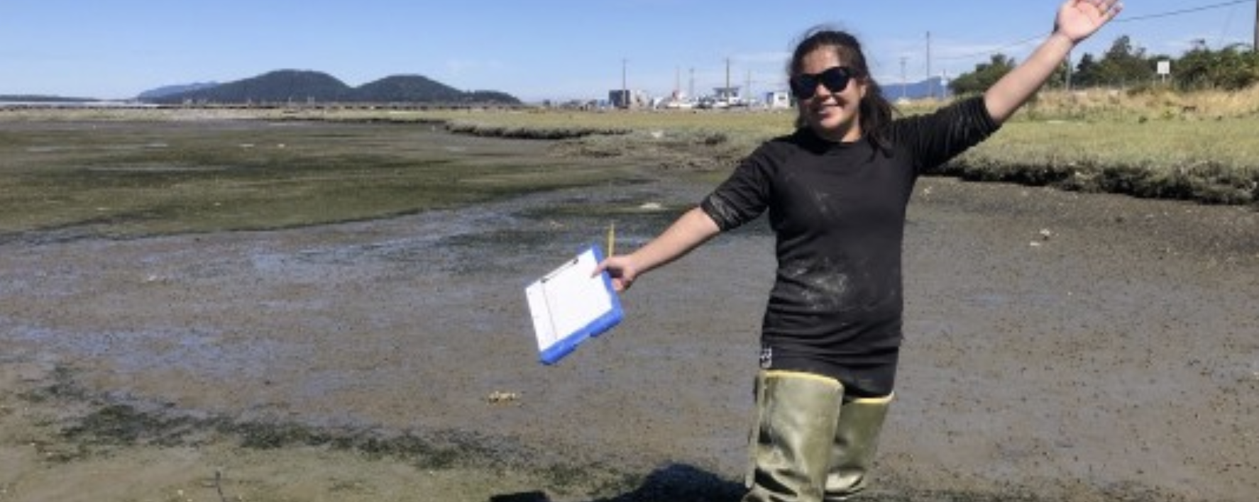 Aina posing to the camera in Fidalgo Bay. Her hands are in the air as she smiles and holds a clipboard 