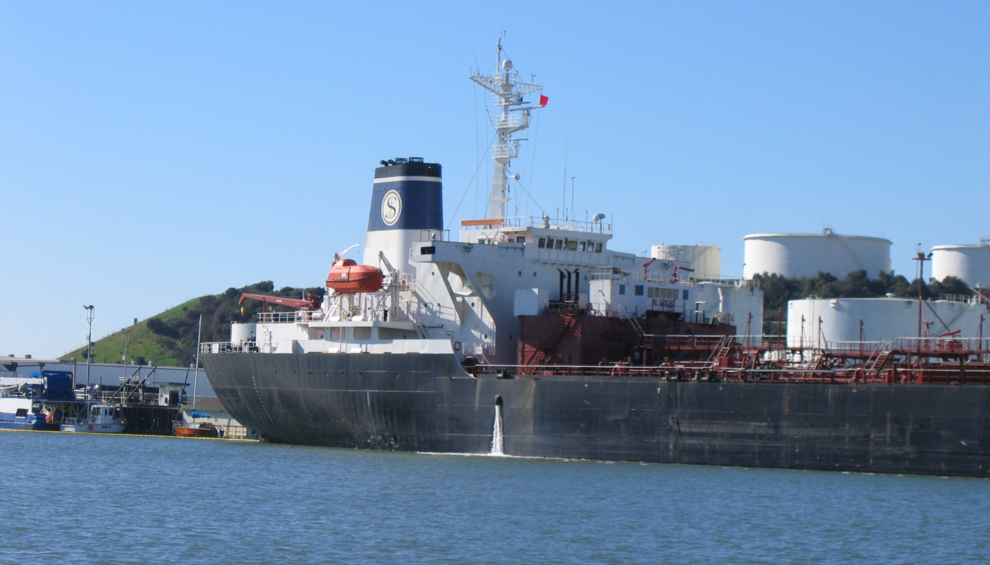 A large cargo ship is in the distance, with a plume of water discharging from its side in roughly the middle of the vessel.