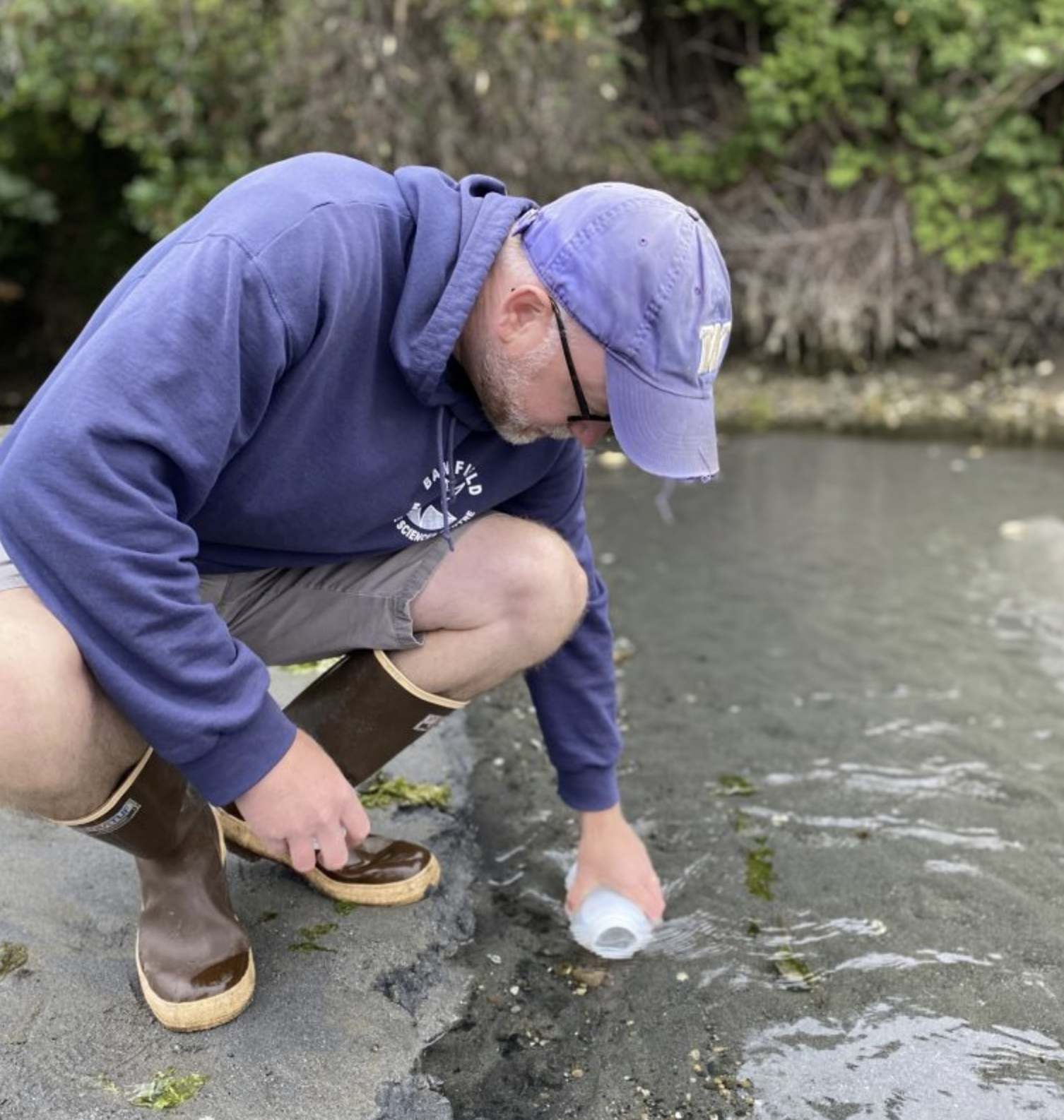 A person leans down to dip a bottle of water in a sandy channel.
