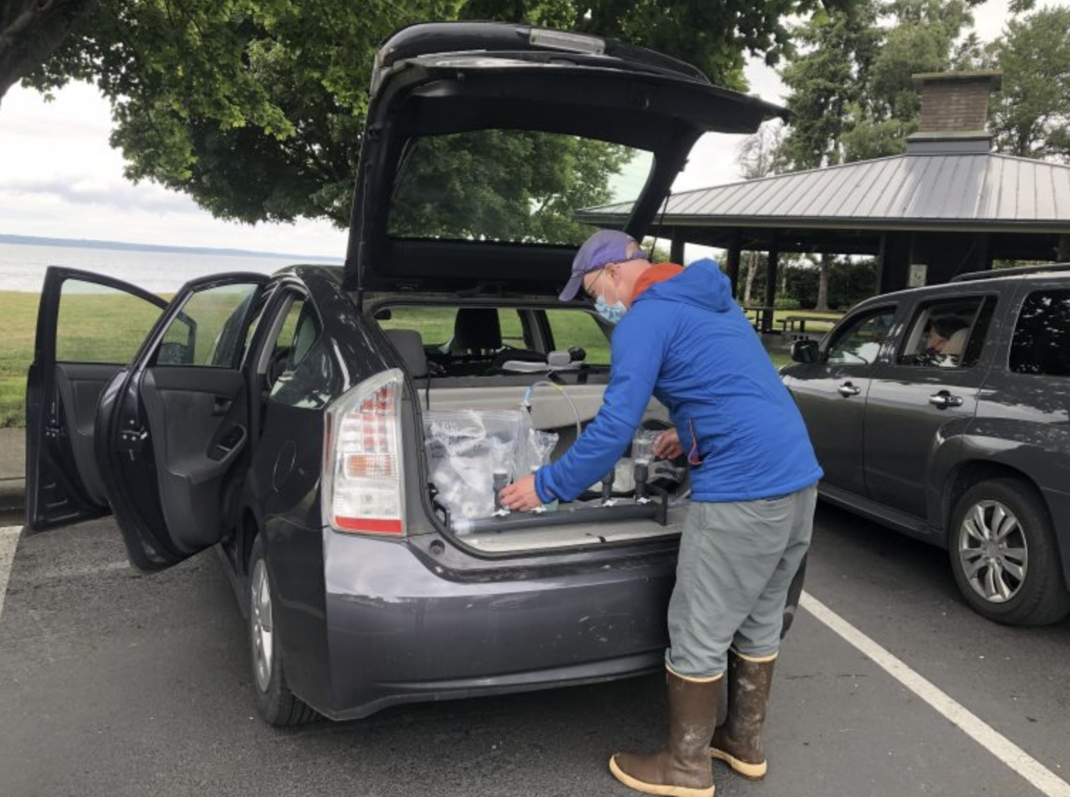 A photo shows a person standing in front of an open tailgate of a Prius. In the trunk is a water filtration set up, with a vacuum pump run by the car's battery, and filtering eDNA water samples.
